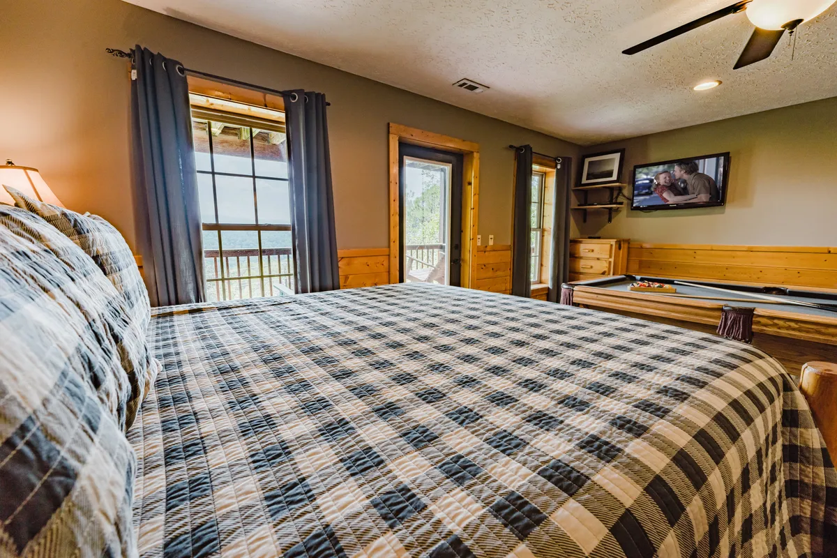 Here we see a serene bedroom with plaid bedding, large windows that frame the mountain view, and a wall-mounted TV. The sunlight pours in, making the space feel bright and welcoming. The arrangement invites relaxation, with the promise of direct access to outdoor vistas and cozy evenings spent lounging or watching TV.