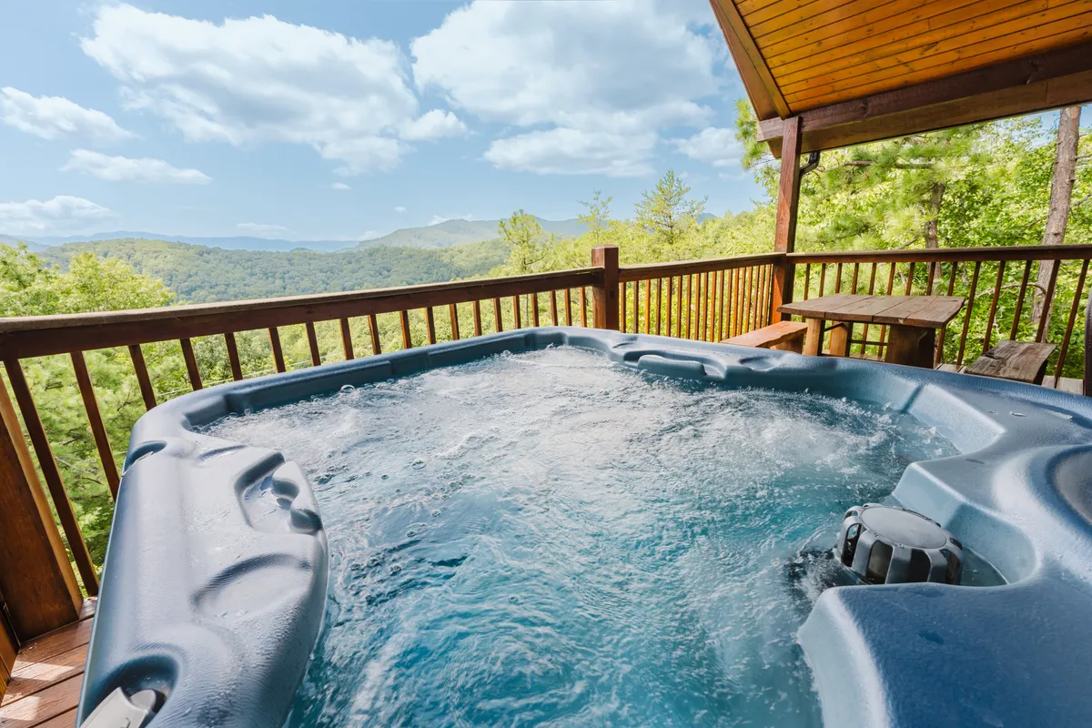 A bubbling hot tub sits on a wooden deck that looks out over expansive mountain scenery. The setting is both private and inviting, ideal for unwinding together as the sun sets or under a canopy of stars. The area also offers space for drinks or light snacks, enhancing the sense of indulgence and relaxation.