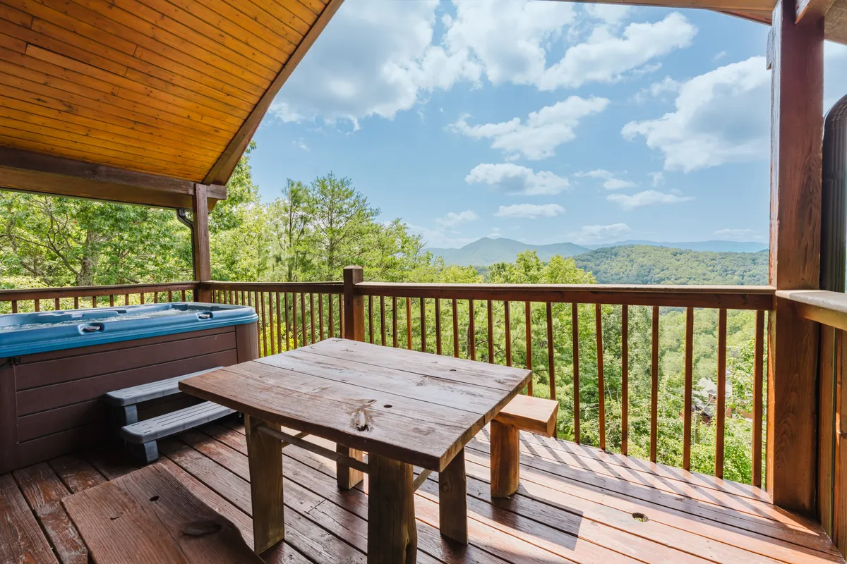 A serene exterior deck scene featuring a wooden table set for outdoor dining, a private hot tub, and a sweeping mountain view beyond the wooden railing. The inviting setup hints at relaxation, memorable meals, and soaking in nature's beauty.