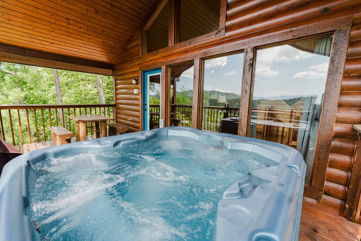 An interior view featuring a hot tub positioned by large windows and wooden beams, offering sweeping mountain or forest views directly from the comfort of the cabin. The scene is tranquil and invites relaxation while staying connected to nature outside.