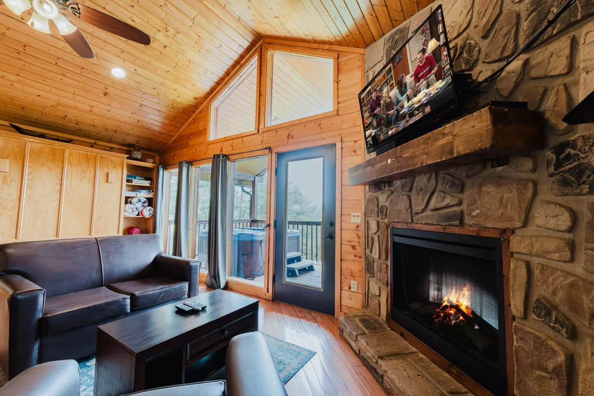 A cozy living area with a stone fireplace, plush sofa, large windows letting in natural light, a television, and a wooden ceiling overhead. The space is arranged for gatherings—perfect for relaxing after a day of adventure, sharing stories, or enjoying a movie night.