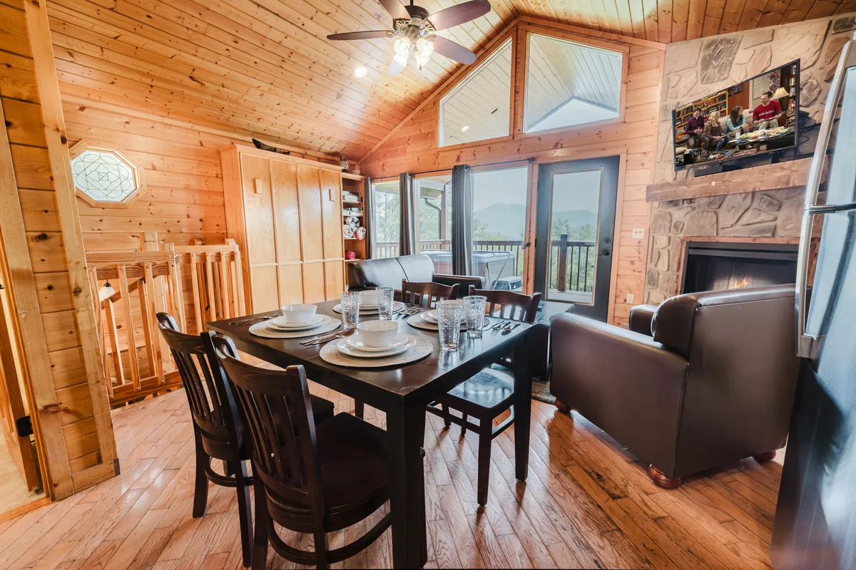 A view of the main living and dining area featuring a dining table set for a meal with large windows offering stunning mountain views. A stone fireplace creates a cozy focal point, while comfortable seating invites relaxation, perfectly capturing the space where guests gather for meals, games, or quiet evenings by the fire.