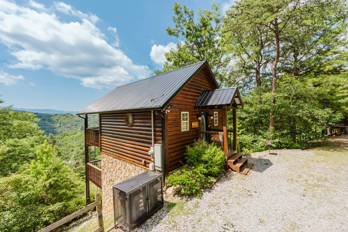 A charming log cabin set amongst lush trees with mountains visible in the background and a gravel pathway leading up to the entrance. The exterior exudes a cozy, inviting vibe, and the natural surroundings hint at outdoor adventures.