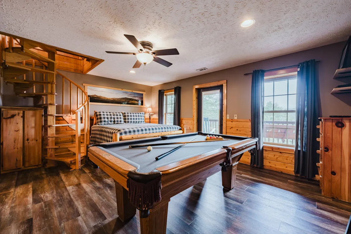 The image shows a sunlit space with a pool table, inviting bed, wooden staircase, and large windows. The natural light streaming in offers a glimpse of the outdoor serenity, while the cozy arrangements make it clear this is a multifunctional space for both restful nights and playful moments. The bed and pool table are central, emphasizing the unique combination of intimacy and entertainment.