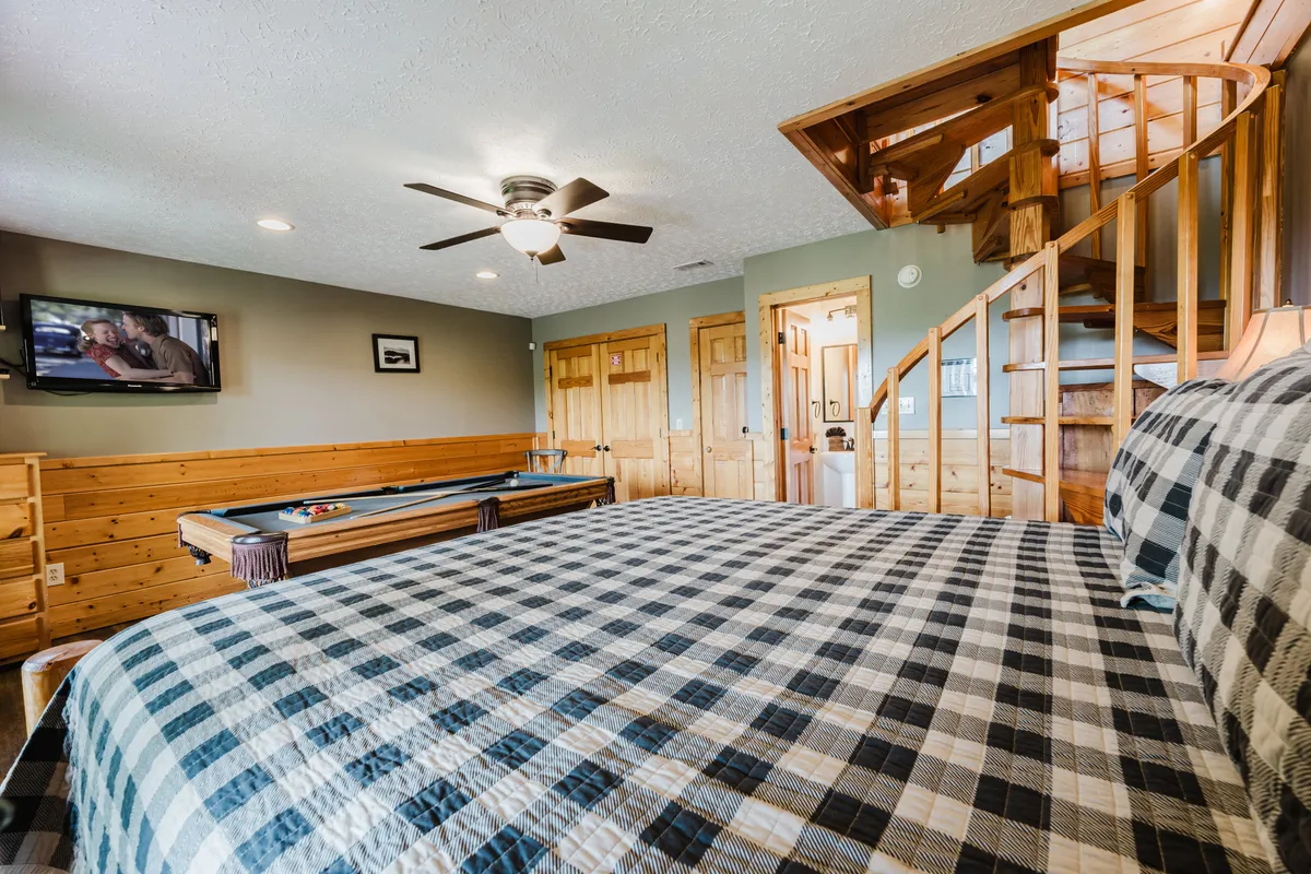This image features a cozy bedroom with a plaid bedspread, a wooden spiral staircase leading down to the space, a pool table, and a wall-mounted television. The decor feels warm and inviting, with wood accents and soft lighting creating a sense of privacy and comfort. The layout visually connects the sleeping area to the game nook, highlighting the suite's playful and intimate design.