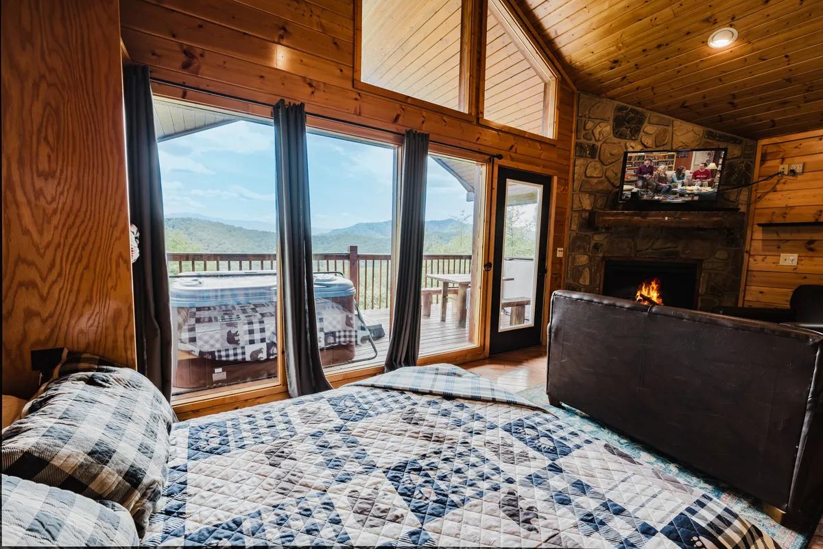 This image captures a spacious living area with large windows offering impressive mountain views, a cozy fireplace, comfortable seating, and a hot tub located just outside on the deck. The scene embodies relaxation after a day of adventure, with visual cues to both indoor comfort and outdoor scenery.
