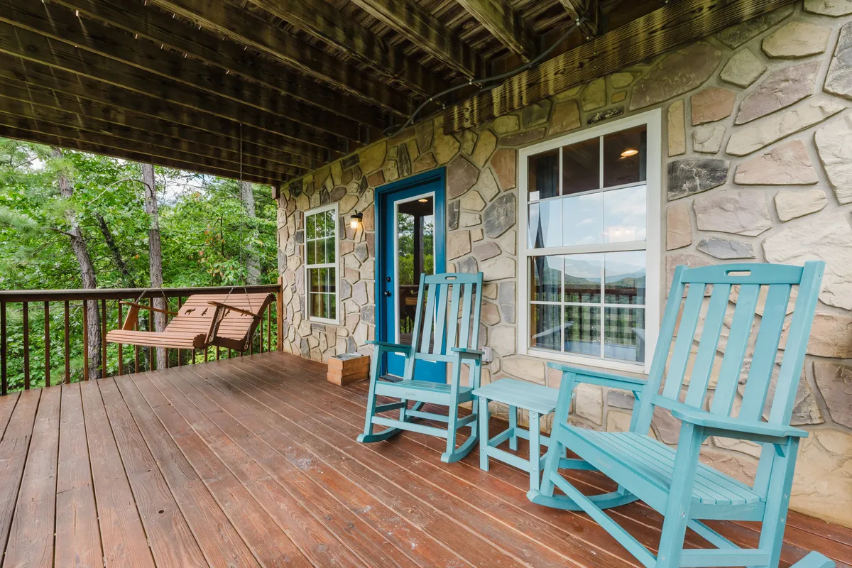 A welcoming exterior scene with a rocky stone wall, blue door, turquoise rocking chairs, and a swing bench on the porch, set against a natural backdrop. The porch overlooks peaceful surroundings, inviting outdoor relaxation and immersion in nature.