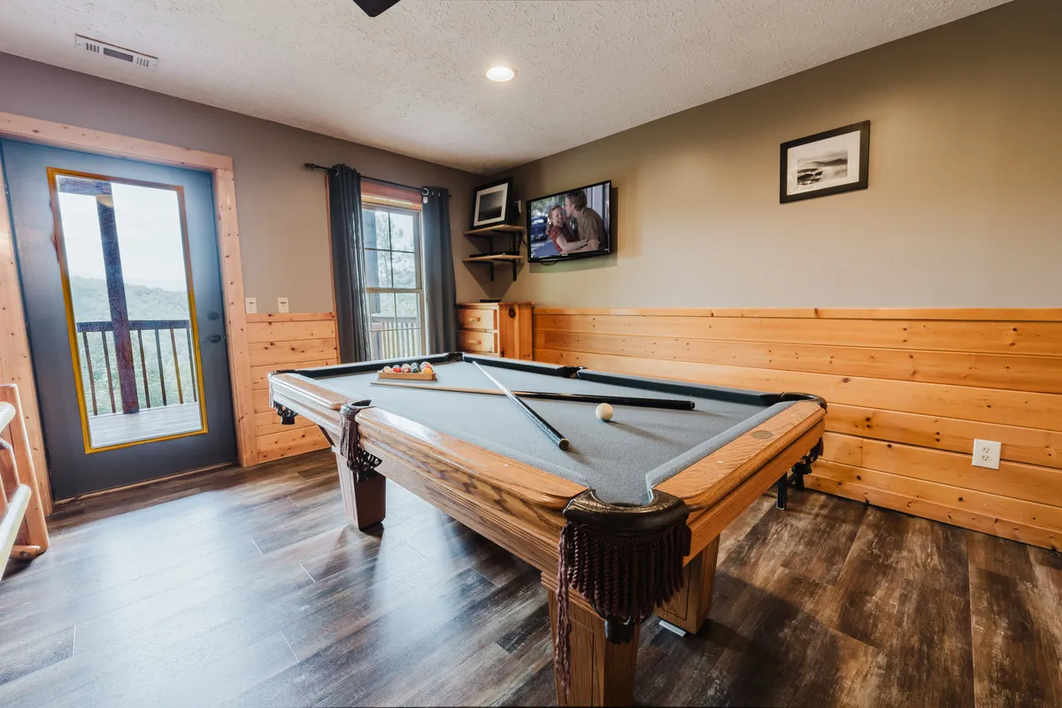 A fun recreation room with a pool table, large window letting in natural light, television, and wood-paneled walls. The space is set up for friendly competition and laid-back evenings, with outside views that hint at the cabin’s scenic location.